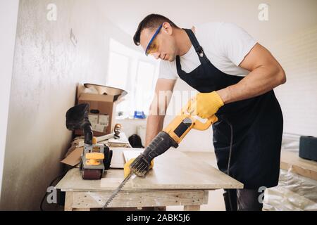 Maintenance man is using a power saw to fix furniture in a room under construction. He is wearing glasses, yellow gloves and  a black apron Stock Photo