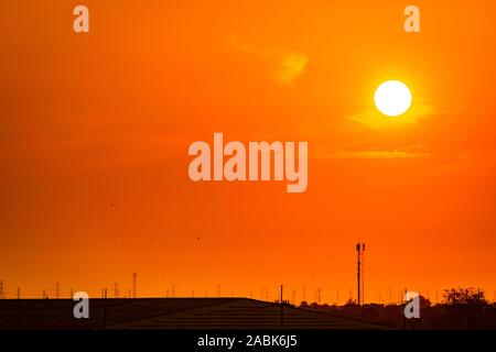 Beautiful orange sunset sky with roof of factory and birds flying near high voltage electric pylons. Evening sunset sky at industrial zone. Power and Stock Photo
