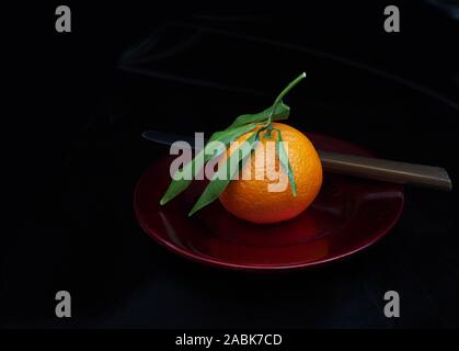 Mandarin type small orange on red plate, with leaves. Whole, unpeeled. With knife, dark background. Stock Photo