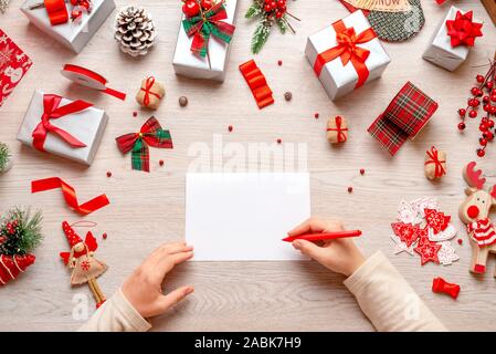Writing Christmas and New Year greetings. Hand holds pen over blank paper. Mockup, copy space. Flat lay, top view composition with loats of gifts and Stock Photo