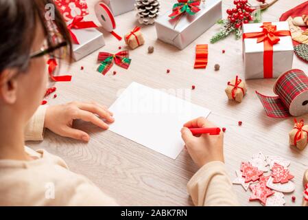 Girl writes Christmas and New Year greetings. Blank paper to add greeting card text. Mockup. A view over shoulder at a table full of gifts and decorat Stock Photo