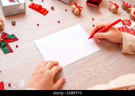 Blank paper and hand with pen surrounded with Christmas gifts and decorations. Concept of writing Christmas and New Year greetings. Close-up. Mockup. Stock Photo