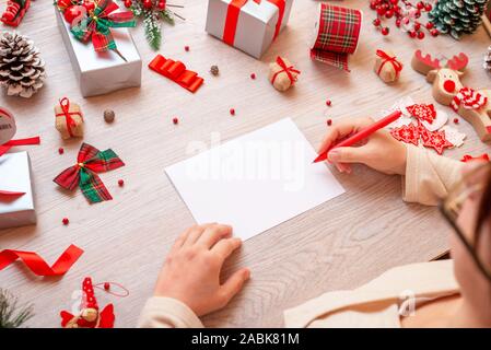 Girl writes on paper surrounded by lots of Christmas gifts and decorations. Paper is blank, to add greeting card text. Stock Photo
