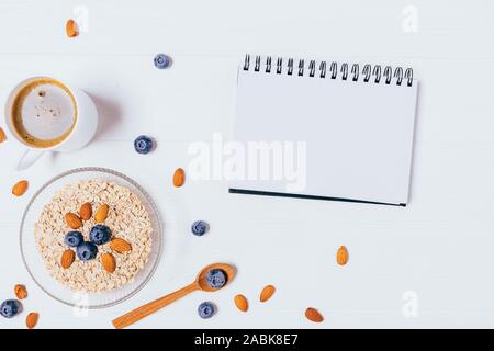 Blank notepad near bowl of oat flakes with blueberries and almonds next to black coffee on white wooden background, top view. Stock Photo