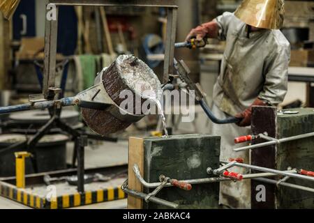 A craftsmen in silver protective clothing performing a metal sand casting technique by pouring molten aluminum silver colored liquid into a mold in a Stock Photo