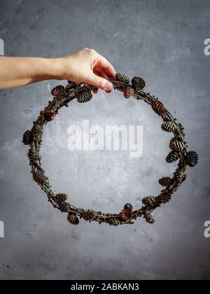 Woman's hand holding a larch wreath with cones as a symbol of natural Christmas on a grey background Stock Photo