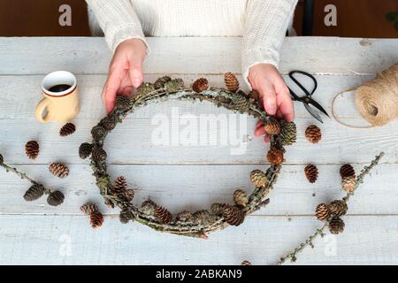Fragment of a woman making a handmade larch wreath with cones as a symbol of natural Christmas on a white wooden table Stock Photo