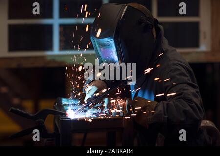 Focused welder designer wearing a helmet and wokring on a project by welding pieces of metal while making welding sparks in his workshop at night Stock Photo