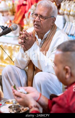 A Hindu priest leads his congregation in morning prayers sitting next to a man holding a lotus flower. In South Richmond Hill, Queens, New York City. Stock Photo