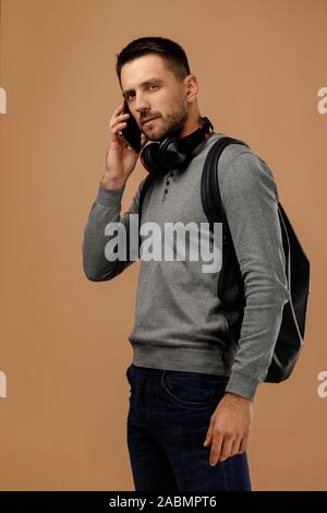 handsome young man with headphones and backpack talking on phone isolated on beige background. Stock Photo
