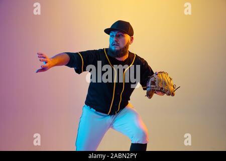 Baseball player, pitcher in a black uniform practicing and training on gradient background in neon light. Young professional sportsman in action and motion. Healthy lifestyle, sport, movement concept. Stock Photo