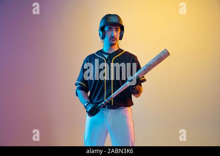 Baseball player, pitcher in a black uniform posing confident on gradient background in neon light. Young professional sportsman in action and motion. Healthy lifestyle, sport, movement concept. Stock Photo