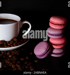 A white cup with black fragrant coffee stands on a black background with coffee zurnes next to colorful french cookies macaroons Stock Photo