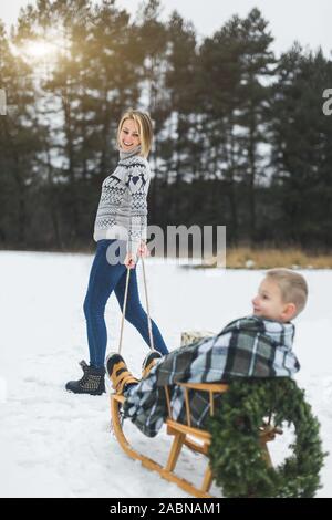 Pretty young mother pulling decorated wooden sled with her son sitting on it in winter snowy day at nature. Stock Photo