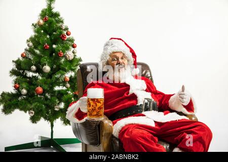 Santa Claus drinking beer near the Christmas tree, congratulating, looks drunk and happy. Caucasian male model in traditional costume. New Year 2020, gifts, holidays, winter mood. Copyspace for your ad. Stock Photo