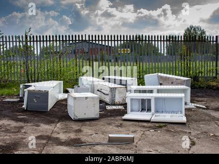 Several discarded fly-tipped fridges and freezers dumped on concrete ground in front of spiked security fence, Clayton, Manchester, UK. Stock Photo