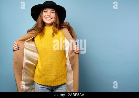 A brunette in a yellow sweater and black hat stands on a blue background, revealing a beige coat to the sides.  Stock Photo