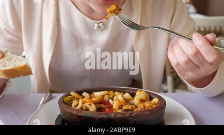 Good served food in restaurant. Unrecognizable woman is eating frying hot shrimps with vegetables served in clay bowl, closeup dish. Takes shrimp out of the boiling oil with a fork. Stock Photo