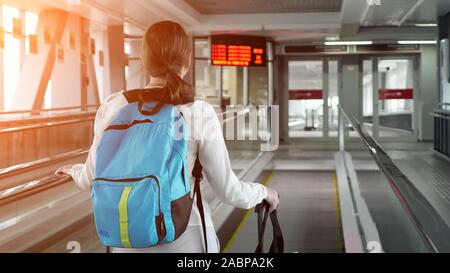 Young woman traveller on escalator with backpack and suitcase in airport terminal, sunlight. She is going to to the airport exit, copy space. Stock Photo