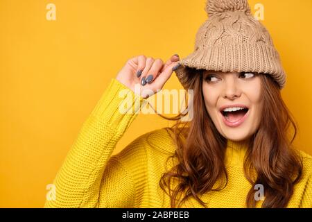 A brunette in a yellow sweater stands on a yellow background and, raising a hat, looks to the side. Stock Photo
