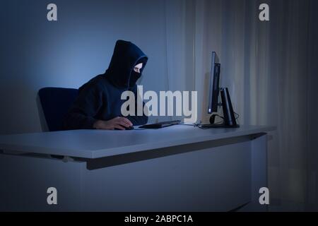 Masked hacker in front at computer into dark room represents the danger of surfing the internet. Internet security concept. Stock Photo