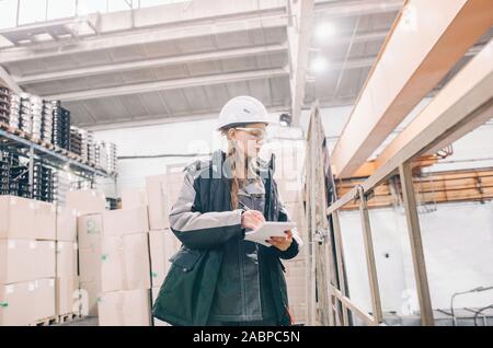 Young female technician in uniform, protective eyeglasses and helmet with notebook. Industrial zone Stock Photo