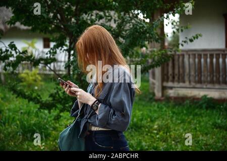 A red-haired girl in a denim shirt stands in profile with head bowed and looks at the phone against the backdrop of a green yard Stock Photo