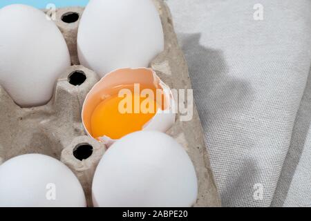Chicken eggs in a white cardboard container lie on a beige wooden kitchen table. One egg is broken. A kitchen towel lies nearby Stock Photo