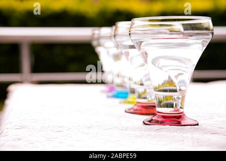 Colorful drinking glasses on white background standing in a row in natural light with green foliage background. red, orange, blue green purple . Stock Photo