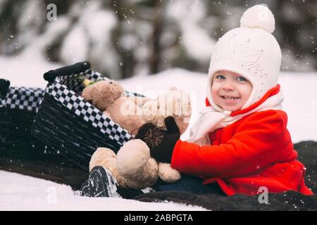 Little girl having fun on winter day. girl in red coat playing with teddy bears on plaid in winter forest. Stock Photo
