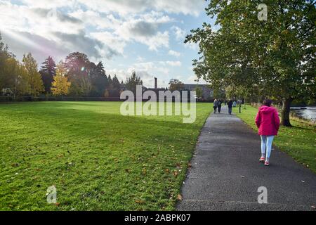 Female in a pink coat walking away from camera on a tree lined riverside path with grassed park to one side. Stock Photo