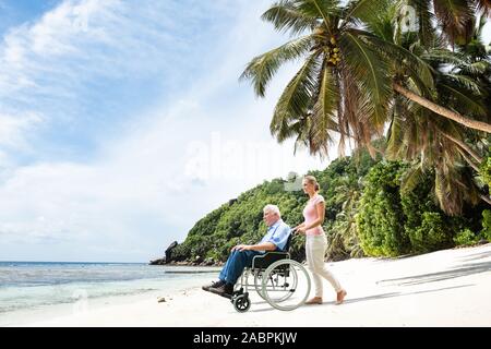 Elder Grandfather In Wheelchair With Granddaughter Walking On Beach Stock Photo