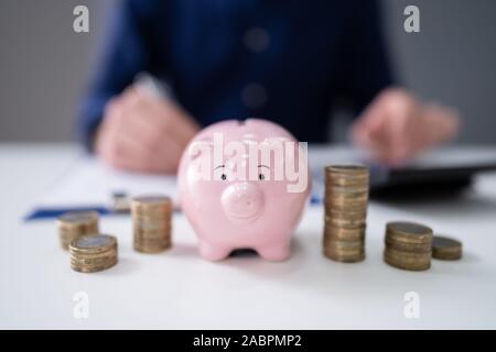 Businessperson's Hand Using Calculator With Stacked Coins And Piggy Bank On Wooden Desk Stock Photo