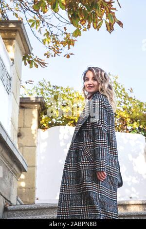 A young Caucasian girl in an autumn checkered coat walks around the city on a sunny day. Looking back towards the camera, climbing up the stairs. Stock Photo