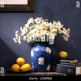 BLUE VASE CONTAINING MICHAELMAS DAISIES & ALSO DECORATED WITH WHITE DAISIES ON A SHELF WITH FRESH LEMONS AND BOOKS INSIDE A HOUSE. Stock Photo