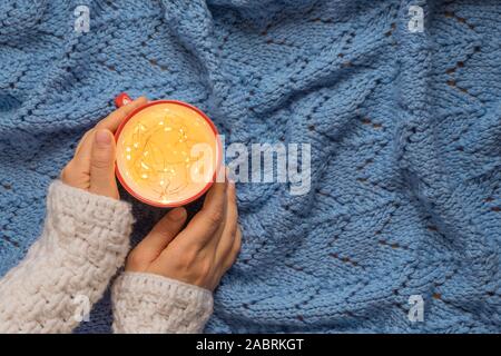Creative Christmas photography concept. Woman hands holding a mug with fairy lights on blue knitted blanket background. Stock Photo