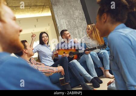Young men and women having fun together indoors drinking beer guy playing guitar others dancing singing participating in party smiling joyful Stock Photo