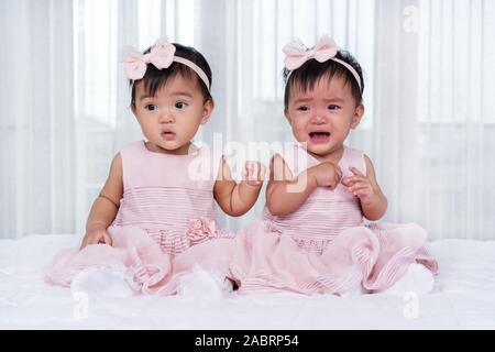 two twin babies in pink dress on a bed, one looking, one crying Stock Photo