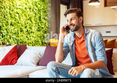 Happy and cheerful young adult man sitting on comfort couch, spending weekend day at home, talking on modern smartphone, looking aside and laughing Stock Photo