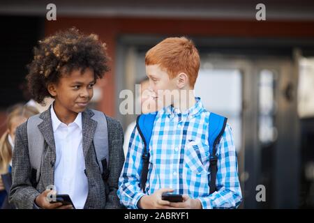 Two schoolboys with mobile phones talking to each other while walking outdoors after school lessons Stock Photo