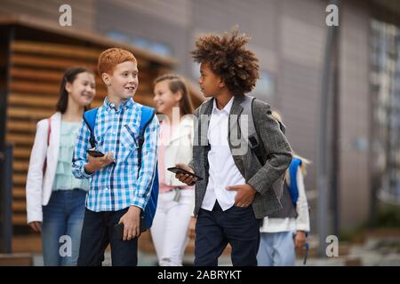 Two schoolboys using their mobile phones and talking to each other while walking along the street with other school children in the background Stock Photo