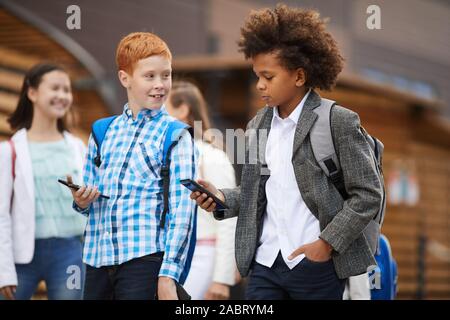 Two friends using their smart phones while walking along the street after school with their classmates in the background Stock Photo