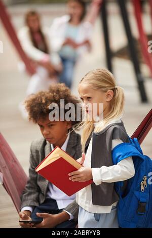 Blond schoolgirl with backpack behind her back holding book standing on the playground with African boy who using mobile phone Stock Photo