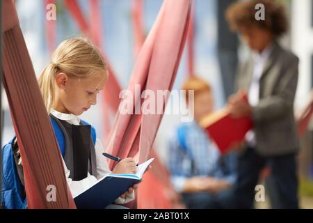 Serious schoolgirl with backpack behind her back making notes in notebook while sitting on the playground with her classmates in the background Stock Photo