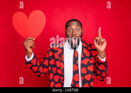 Photo of funny dark skin man cupid hold big paper heart shape raise finger have idea for romantic surprise open mouth wear hearts pattern suit shirt Stock Photo