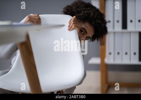 Frightened Young Businessman Hiding Behind Chair At Workplace Stock Photo