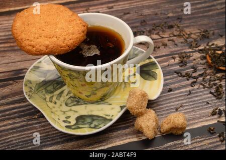 A Cup of black tea, tea leaves, pieces of brown sugar, oatmeal cookies on a wooden background. Close up Stock Photo