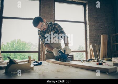Photo of pensive man wearing checkered shirt eye glasses protecting him from sawdust working by using sander Stock Photo