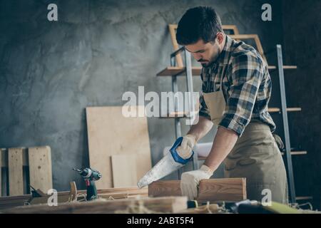 Side profile photo of serious confident focused man cutting off unneeded pieces of wooden block using saw Stock Photo