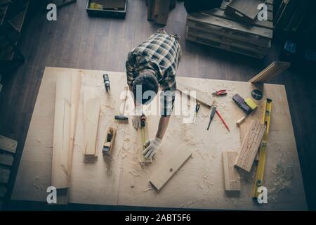 Top high above angle view photo of man measuring the length of wooden piece in checkered shirt surrounded with instruments indoors Stock Photo